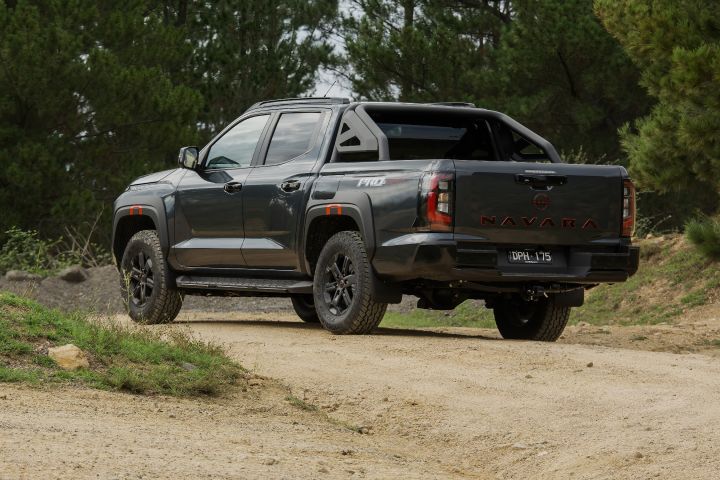 2026 Nissan Navara PRO-4X finished in Boulder Grey shown wide from the rear three‑quarter on a winding trail with side steps, black alloys, sports bar and NAVARA tailgate script.