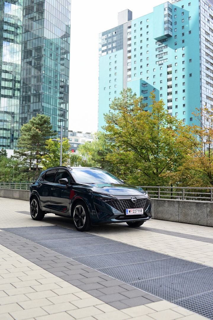 Hochformatiges Bild eines dunkelgrünen Nissan Qashqai e‑Power, der vor zwei Hochhäusern steht