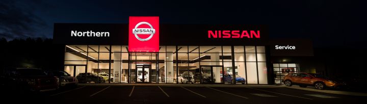 Vue nocturne d'un concessionnaire Nissan avec signalisation lumineuse, présentant des voitures dans la salle d'exposition et une zone de service.