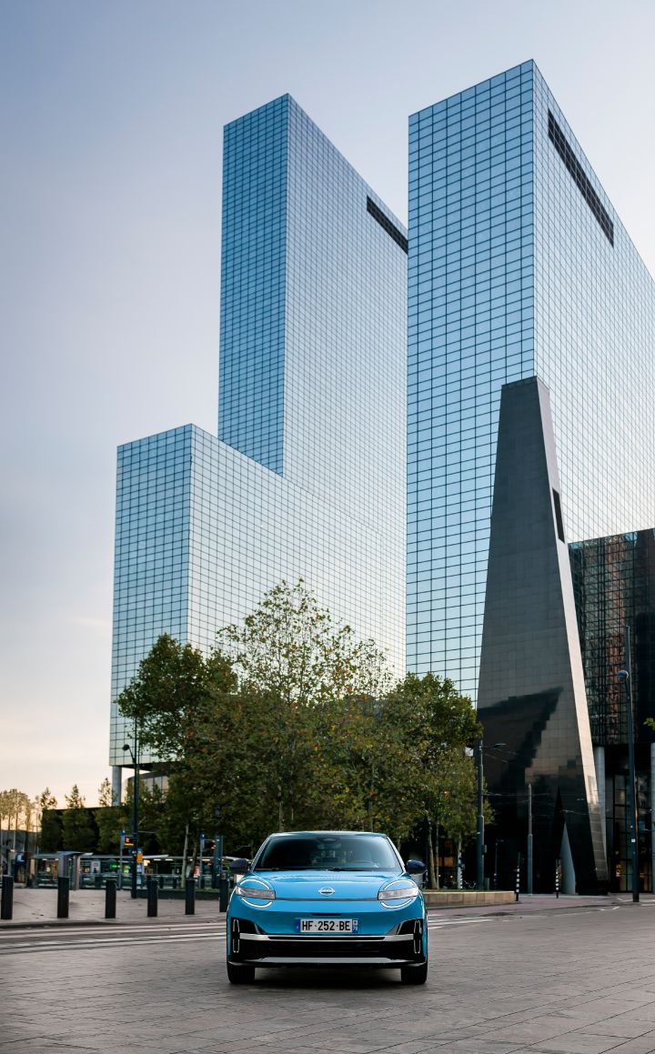 Frontalansicht eines blauen Nissan Micra, der in Rotterdam vor zwei Hochhäusern mit spiegelnder Glasfassade steht.
