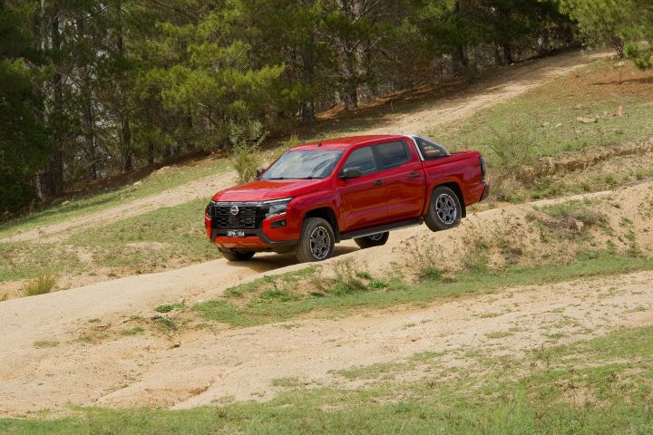 A red 2026 Nissan Navara ST‑X driving over an uneven sandy section of an off‑road track bordered by trees.