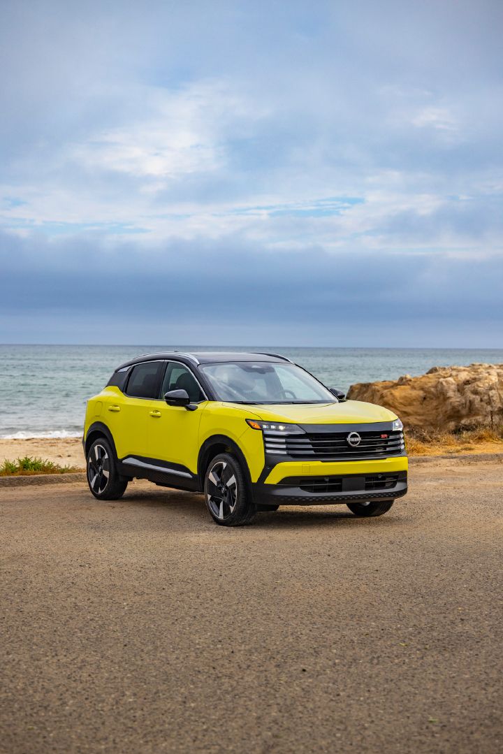 Front right 3/4 view of a neon yellow 2025 Nissan Kicks parked on a gravel lot overlooking the ocean and large mass of clouds.