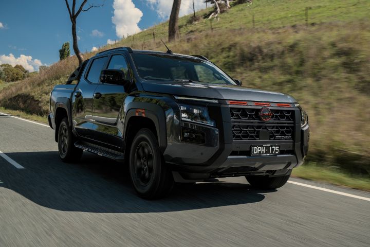 2026 Nissan Navara PRO-4X finished in Boulder Grey shown in a front-quarter rolling shot beside a grassy embankment with dark wheels, sports bar and red grille bar.