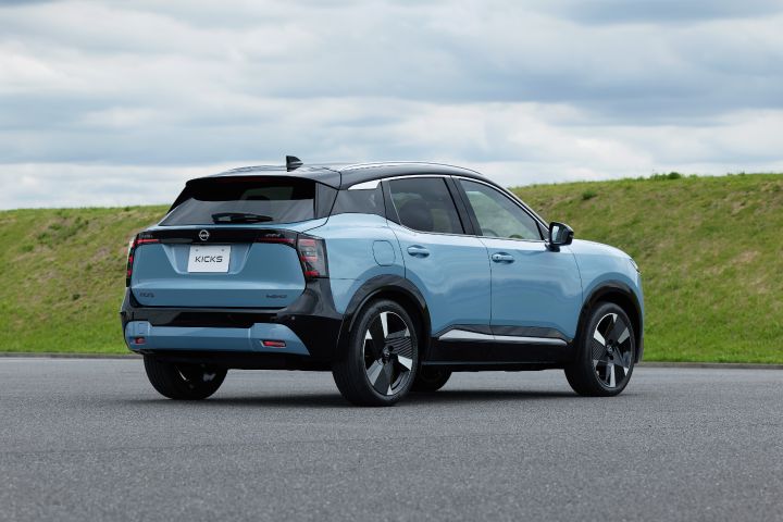 青みがかったキックスの後方斜めビュー。車両は右向きに進んでおり、背景には舗装路と草地の斜面が見える静止に近い構図。