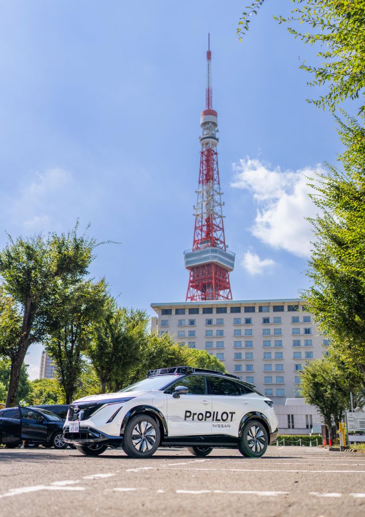 Ein weißer Nissan Ariya ist mit der Front nach links geparkt, an seiner Seite ist das Wort „ProPILOT” zu sehen. Im Hintergrund ragen ein Hochhaus und der Tokyo Tower vor blauem Himmel empor, umgeben von Bäumen