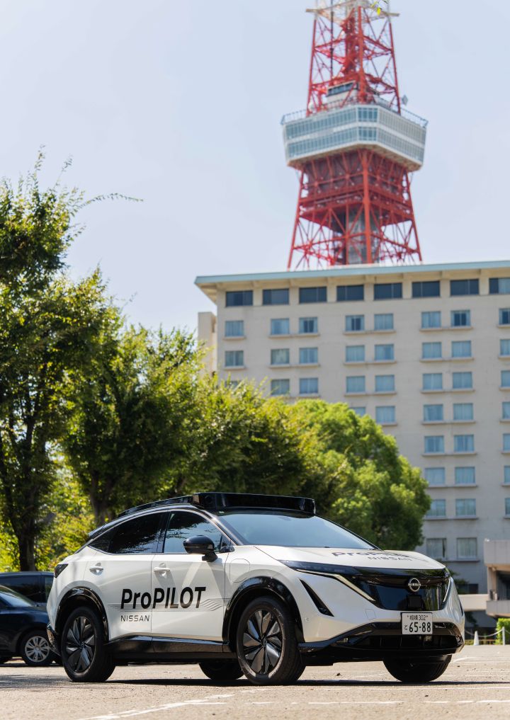 Ein weißer Nissan Ariya ist mit der Front nach links geparkt, an seiner Seite ist das „ProPILOT”-Logo zu sehen. Im Hintergrund sind üppig grüne Bäume und der Tokyo Tower zu sehen.