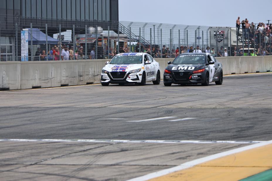 Two Nissan Sentra Cup race cars driving side by side on the race track with spectators in the background