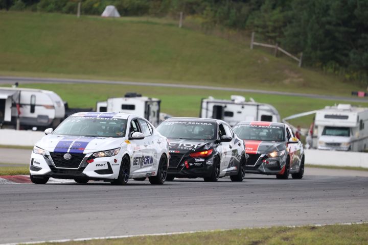 Three Nissan Sentra Cup race cars numbered 21, 17, and 1 compete on an asphalt track; spectators’ vehicles are parked in the background.