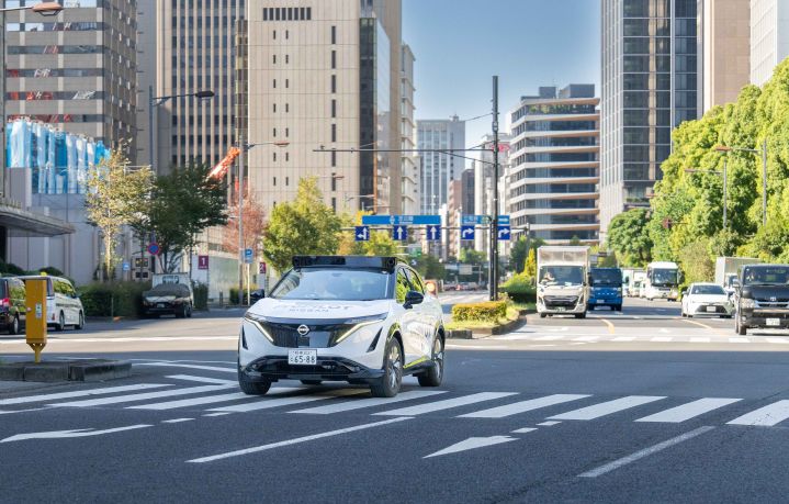 Ein weißer Nissan Ariya fährt über eine Kreuzung in der Stadt. Im Hintergrund sind hohe Gebäude und grüne Bäume zu sehen, außerdem sind weitere Fahrzeuge auf der Straße unterwegs. Der Himmel ist klar und blau.