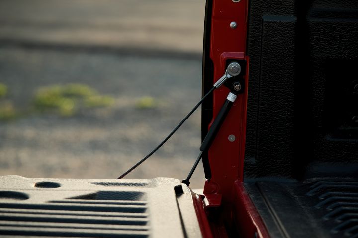 Close-up of the open tailgate support area on a 2026 Nissan Navara ST‑X in Outback Red, showing the retaining cable, damper strut, and textured black bedliner along the inner tray edge.