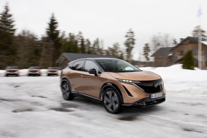 Dynamische Aufnahme vom Elektroauto Nissan Ariya e‑4ORCE in Akatsuki Copper mit Dach und Außenspiegel in Pearl Black, das auf schneebedeckter Fahrbahn schräg vorbeifährt