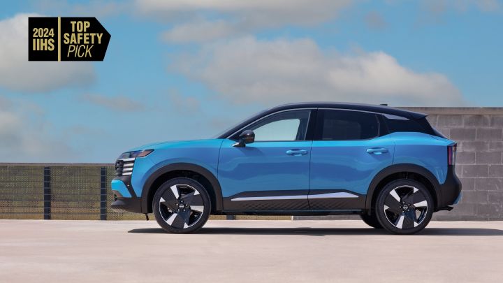 Blue Nissan Kicks facing left with a blue sky and clouds in the background parked on a concrete surface with a wall behind it