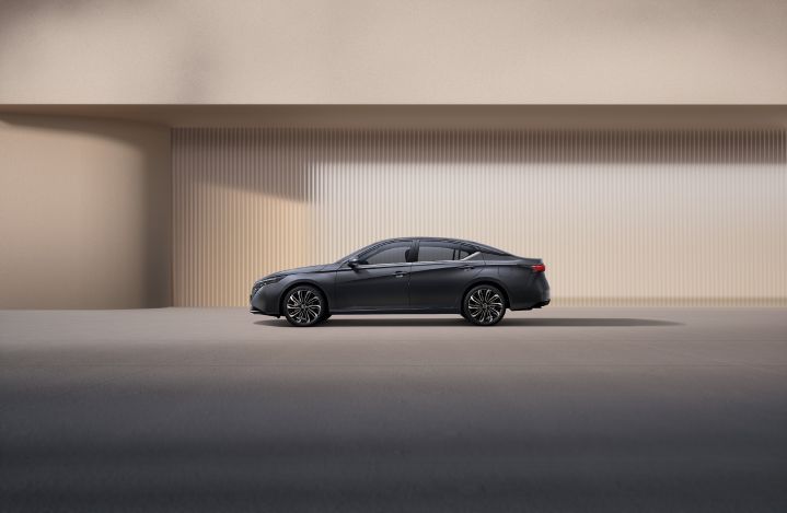 A black Teana parked in a minimal outdoor setting, facing forward. The grille and headlights are prominently visible. The background features light beige and gray walls with soft lighting.
