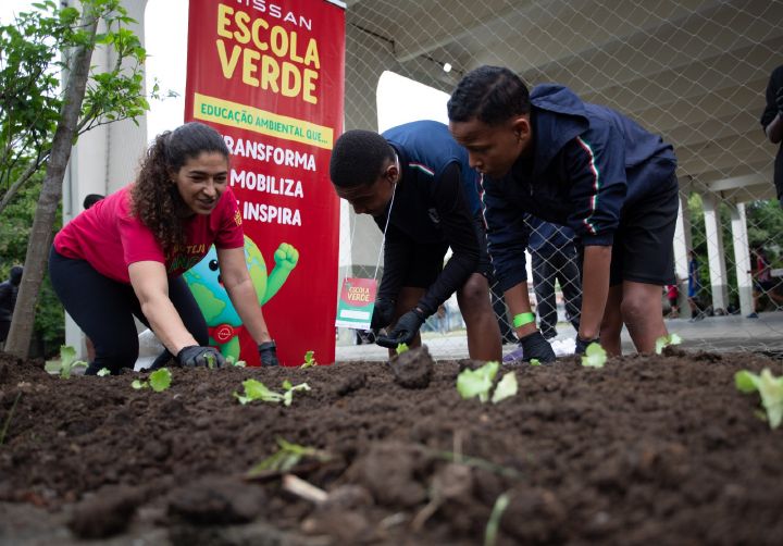 Professora de camiseta vermelha e dois alunos com uniformes azuis-marinho estão agachados cuidando de uma horta. Ao fundo, uma faixa vermelha traz o logo da Nissan em branco e o da Escola Verde em amarelo. Abaixo deles, há as frases “Educação ambientam que... Transforma, Mobiliza e Ispira”