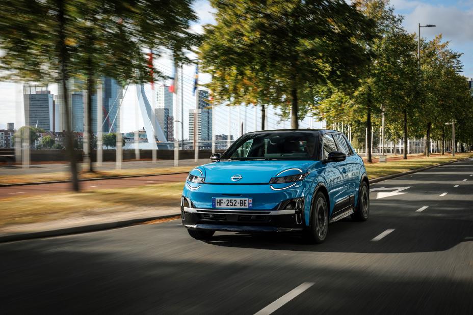 Vue de trois quarts avant d'une Nissan Micra bleue roulant dans le centre-ville de Rotterdam, divers bâtiments de la ville scintillent à travers les arbres derrière elle.