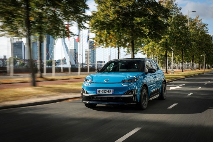 3/4-Frontansicht eines blauen Nissan Micra, der durch die Rotterdamer Innenstadt fährt, durch die Bäume dahinter schimmern diverse Gebäude der Stadt