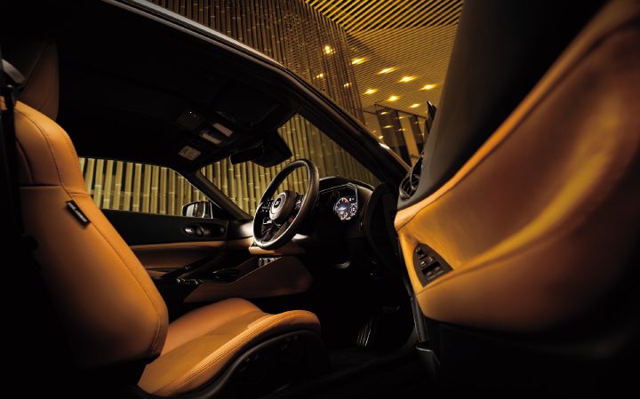 Interior view of a Fairlady Z with a tan cabin, photographed diagonally through an open door from the rear side of the cockpit. Tan sport seats occupy the foreground, while the right-hand-drive steering wheel, instrument cluster, and center console are visible toward the front. The combination of tan leather trim and a black dashboard creates a premium appearance. In the background, a warmly lit interior space with wood-slat wall panels is visible, providing an upscale setting that highlights the sporty and refined character of the Fairlady Z interior.
