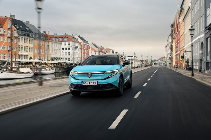 Türkisfarbener Nissan LEAF, der auf einer Stadtstraße durch Kopenhagen fährt, die von einem malerischen Kanal und typisch dänischen farbigen Gebäuden gesäumt ist, fotografiert von vorne links.