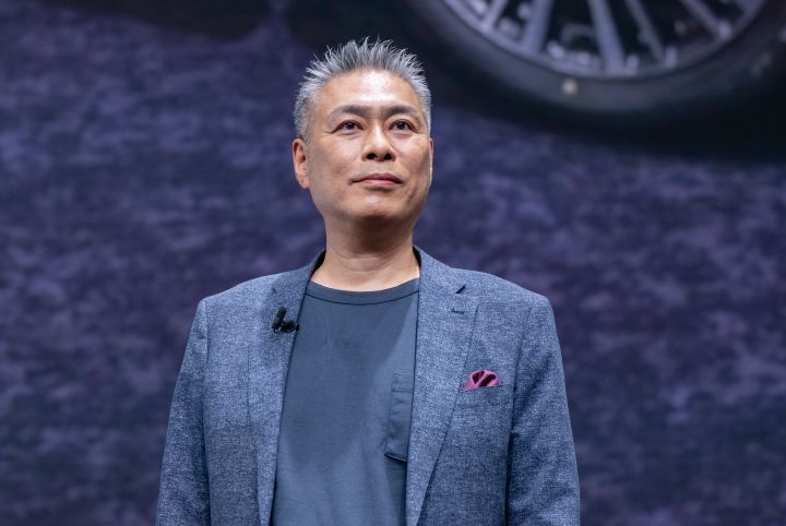 発表会場のステージで登壇する一野CVEの上半身を捉えた写真。一野CVEはブルーのジャケットとダークカラーのTシャツを着用し、正面を向いて立っている。背景には大型スクリーンが広がり、新型エルグランドに関連する車両画像の一部としてホイール付近が大きく映し出されている。青みがかったグレーを基調とした映像と照明が会場全体を包み、プレゼンテーション中の一野CVEを印象的に映し出している。新型エルグランドの技術やデザインを紹介する発表シーンの一場面。