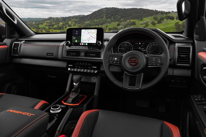 2026 Nissan Navara PRO-4X finished in Boulder Grey presented in a wide right-hand-drive cockpit view with navigation screen, steering wheel and red-accented seats.