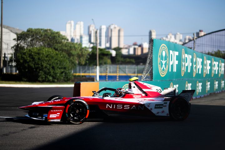 Seitliche Fahraufnahme von Norman Nato im Nissan Formel E Rennwagen mit der Startnummer 23 mit grünen Werbetafeln auf der rechten Seite des Fotos und Bäumen im Hintergrund.