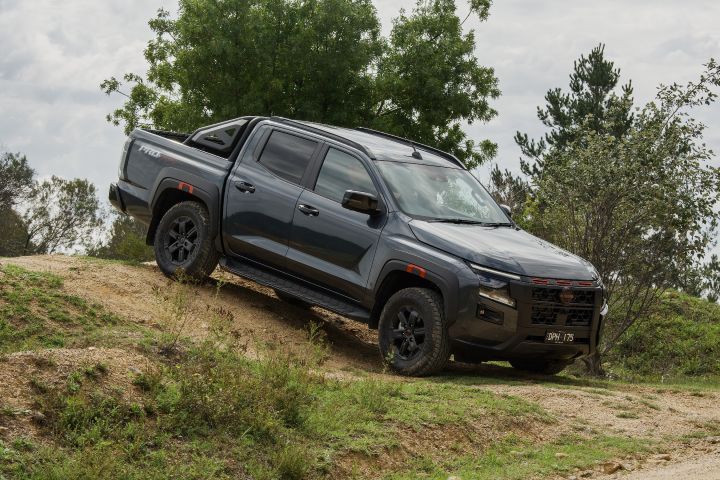 2026 Nissan Navara PRO-4X finished in Boulder Grey descending a small bank side‑on with suspension compression and orange arch accents.