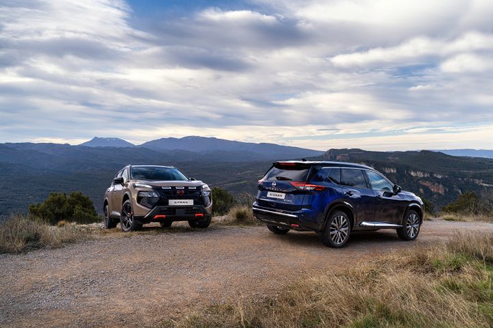 Neuer 2026 Nissan X‑Trail in der Karosseriefarbe „Baja Storm”, geparkt auf einem Feldweg mit Blick auf eine bergige Landschaft und bewölkten Himmel. Das Fahrzeug verfügt über schwarze Felgen und ein schwarzes Dach und steht in der Nähe von Bäumen mit weitläufigem Tal und Hügeln im Hintergrund.