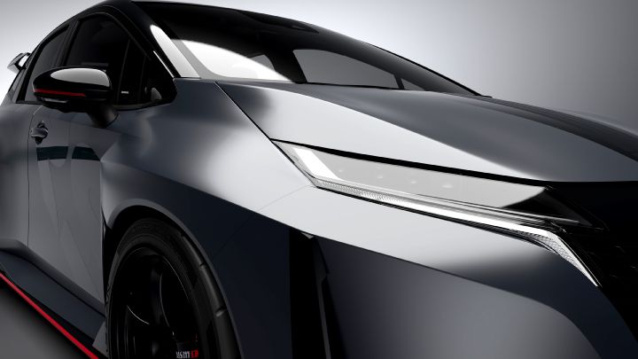 A close-up studio shot of the front-left section of the Aura Nismo RS Concept, a silver and black two-tone sports car. The image highlights the sharp LED headlight and smooth body contours, with a red accent line visible along the lower edge. The black wheel and partially visible red brake caliper add to the sporty look. The background is a simple gray, emphasizing the car's glossy finish and sleek design