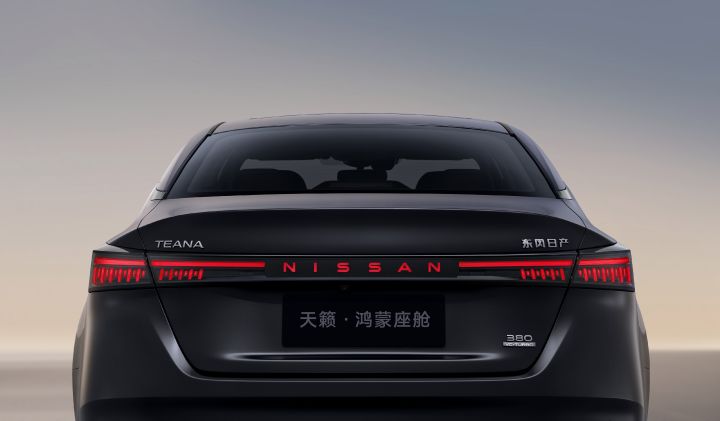 A dark gray new Teana sedan parked on a wide paved surface, viewed directly from the rear. The design features a horizontal tail light strip and a prominent “NISSAN” logo at the center. The background is minimalistic, with a soft gradient sky and no visible buildings or urban elements. The overall tone is subdued, emphasizing the car’s rear styling.