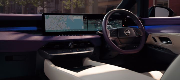Interior cockpit view of the All-new ELGRAND, photographed from the front passenger side. The cabin features a premium two-tone ivory and dark color scheme, highlighted by a wide integrated display stretching across the dashboard. The display shows navigation information and a digital instrument cluster, while a multifunction steering wheel is positioned on the right side. A row of control switches and an electronic gear selector are located at the center of the dashboard, with additional storage space below. Blue ambient lighting runs across the instrument panel and door trim, creating a modern and sophisticated atmosphere. Outside the windshield, trees and building facades are visible, placing the vehicle in an urban setting.