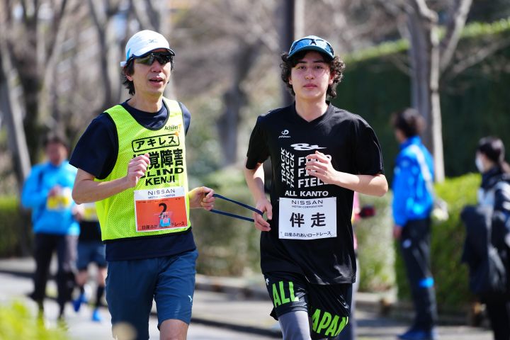 黒いシャツに黒いサングラスを身に着けた男性と、黒いシャツを着た伴走者の男性が走る様子