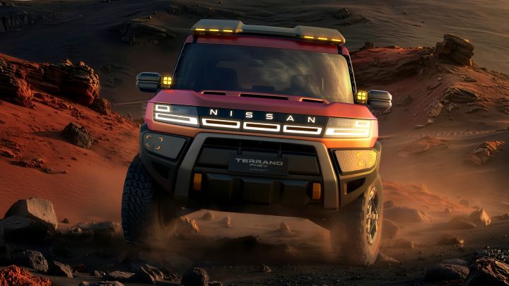 A red Terrano PHEV Concept driving toward the viewer across a rugged desert landscape at sunset. Rocks and sand fill the foreground, with distant mountains and warm evening light in the background.