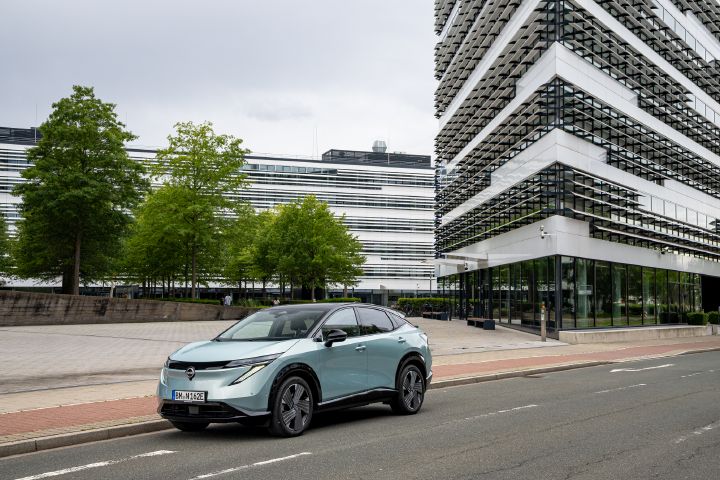 Blick von schräg vorne auf einen hellblauen Nissan Ariya, der am Straßenrand steht mit zwei modernen Bürogebäuden im Hintergrund