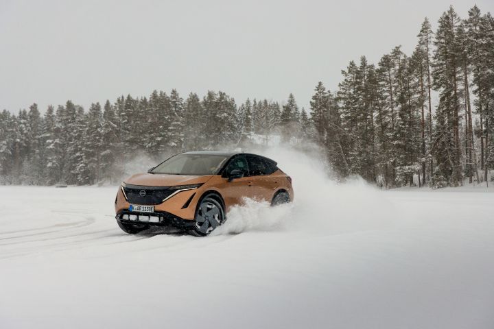 Elektroauto Nissan Ariya e‑4ORCE in Akatsuki Copper mit Dach und Außenspiegel in Pearl Black fährt auf Eissee mit schneebedeckten Bäumen im Hintergrund, 3/4 Front