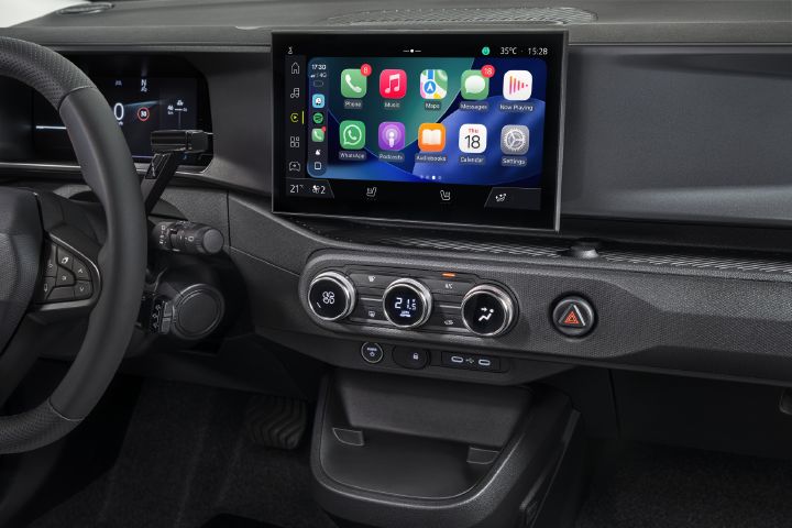 Photograph of a modern vehicle dashboard showing Apple Carplay, steering wheel, climate controls, hazard button, and storage compartment. Center touchscreen displays media tiles and menu labels, while rotary climate dials and illuminated controls organize cabin functions