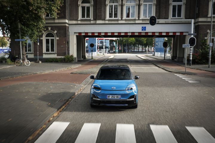 Blue all-new MICRA driving through a city street with a bridge in the background.