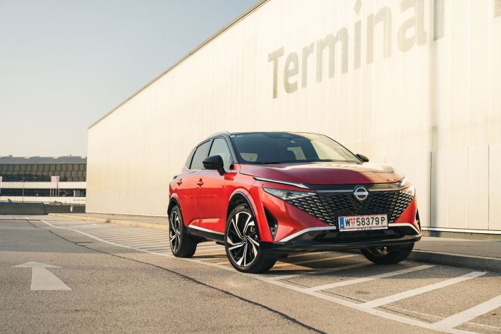 Ein Nissan Qashqai in Fuji Sunset Red steht vor einem fensterlosen Flughafengebäude