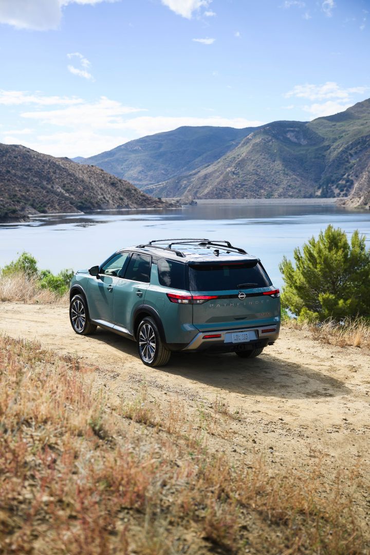 Left 3/4 rear view of the 2026 Nissan Pathfinder parked on a dirt path overlooking tree-covered mountains and water.