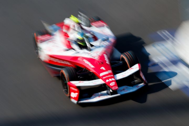 Eine Draufsicht auf einen unscharfen rot-weißen Formel-E-Rennwagen, der schnell auf einer Rennstrecke in Richtung rechts unten fährt. Im Cockpit ist Oliver Rowland zu sehen, während die Fahrbahn im Hintergrund durch die Bewegung unscharf erscheint.