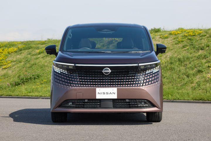 新型エルグランドが、正面からのビューで静止している。車両はカメラに向かってまっすぐ配置されており、大型のブラックグリルと幾何学的なパターン、左右に伸びるLEDヘッドライト、中央の「NISSAN」エンブレムが強調されている。背景には緑の草地と黄色い花が広がる土手があり、上空には淡い青空が広がる明るい日中の屋外シーンで、手前には灰色の舗装路面が見える。