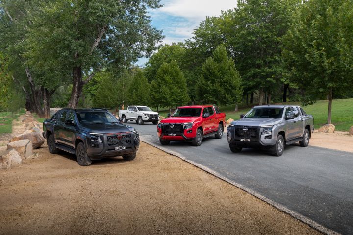 Four Nissan Navara variants positioned across a park setting with gravel and paved surfaces: a Navara PRO-4X in Boulder Grey at left, a Navara ST-X in Outback Red in the middle, a Navara ST in Summit Silver at right, and a Navara SL in Alpine White in the background beneath leafy trees. Landscape orientation