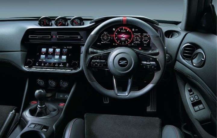 Cockpit view of a Fairlady Z NISMO, photographed from the center rear of the cabin looking forward. The image features a right-hand-drive layout with the steering wheel, digital instrument cluster, and central infotainment display clearly visible. The steering wheel includes a red center marker and red stitching, while the instrument cluster displays a NISMO-specific red-themed graphic design. Three auxiliary gauges are positioned on top of the dashboard, and a six-speed manual transmission shifter is visible on the center console. The predominantly black interior is accented with red highlights, emphasizing the performance-focused character and driver-oriented design of the Fairlady Z NISMO.