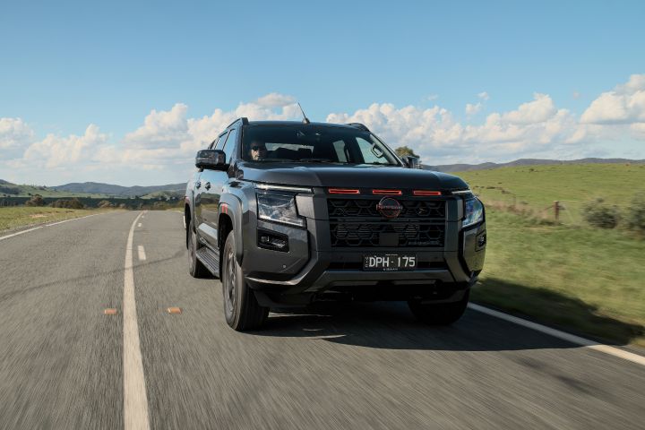 2026 Nissan Navara PRO-4X finished in Boulder Grey depicted in a near-side front rolling shot on open country tarmac with pasture fencing and layered hills.