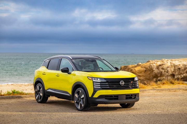 Front right 3/4 view of a neon yellow 2025 Nissan Kicks parked on a gravel lot overlooking the ocean, brush and rocks are behind.