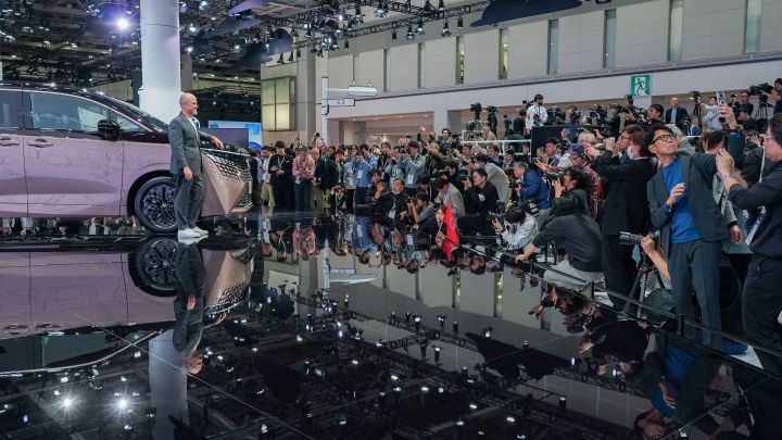 On the stage, the all-new Elgrand in Fuji Dawn and Shigoku two-tone colors is positioned diagonally, facing slightly to the left. Next to the vehicle standsCEO Ivan Espinosa in a suit, delivering a presentation. The background shows a white exhibition setting, while in the foreground, members of the press are gathered, taking photos with cameras.