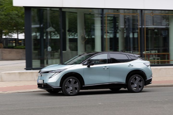 Ein hellblauer Nissan Ariya fährt vor einem modernen Gebäude mit gläserner Fassade über eine Straße, 7/8 Frontaufnahme