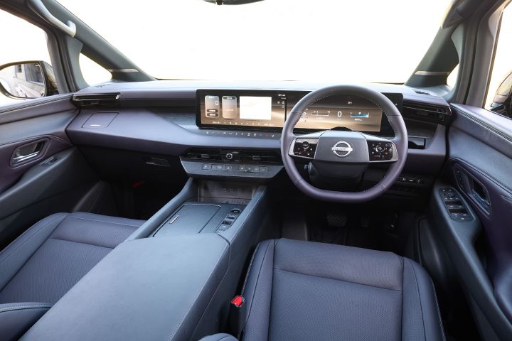 新型エルグランドの運転席周辺のインテリアを撮影した広角ビュー。右側にステアリングホイールが配置された右ハンドル仕様で、中央には横長の大型ディスプレイがあり、デジタルメーターとインフォテインメント画面が一体化している。ダッシュボードとセンターコンソールはダークトーンで統一され、フラットで広がりのあるデザインが特徴。下部には電子シフトボタンや収納スペースが配置され、シートはブラックのレザー調素材。フロントウインドウの外は明るい白い光で、屋外の強い日差しが差し込む室内空間となっている。
