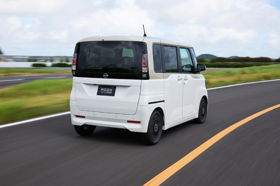 A white, new Nissan Roox kei car is driving on a paved road with a yellow line. The car is viewed from the rear and appears to be in motion. The background includes green grass and distant buildings under a partly cloudy sky.