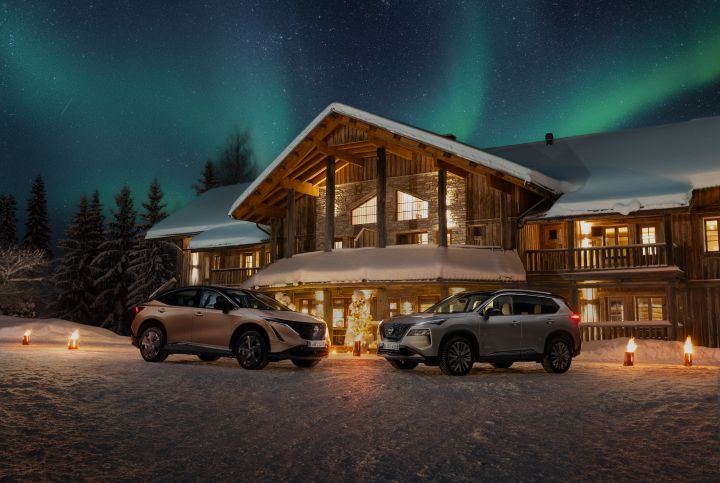 Elektroauto Nissan Ariya e‑4ORCE in Akatsuki Copper mit Dach und Außenspiegel in Pearl Black und silberner Nissan X‑Trail e‑Power e‑4ORCE stehen nebeneinander im Schnee vor verschneitem Holzhaus und Wald, am Nachthimmel leuchten grün die Polarlichter