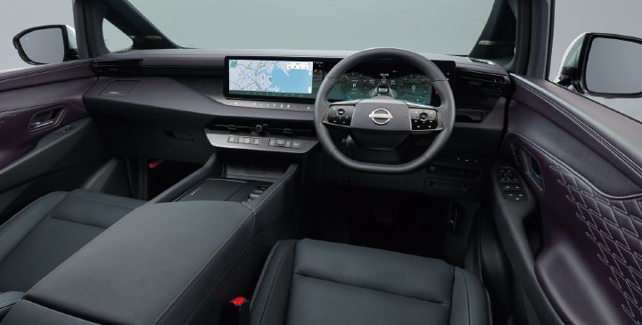 エルグランド AUTECH LINE e‑4ORCE G-specの運転席まわりを、後席中央付近から前方に向かって撮影したインテリア画像。画面中央にはブラックのステアリングホイールが配置され、その奥には横長の大型ディスプレイが広がっている。ディスプレイ左側にはナビゲーション地図、右側にはデジタルメーターや車両情報画面が表示されている。
インストルメントパネルは濃いパープルを基調とし、上質なデザインを採用している。センターコンソールは前席から後方へと続く大型アームレスト形状となっており、落ち着いた高級感を演出している。ダッシュボード中央には空調吹き出し口やシフト関連スイッチ類が整然と配置されている。
左右のドアトリムには立体的なダイヤ柄のキルティング加工が施され、レザー調素材と繊細なステッチが高級感を引き立てている。前席シートはいずれも前方を向いて配置されている。